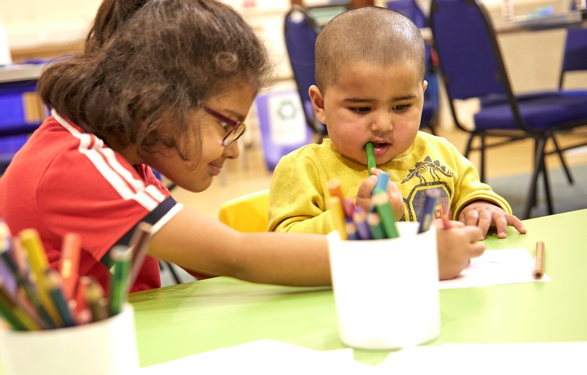 Colour photograph of two children sat at a table colouring a picture.