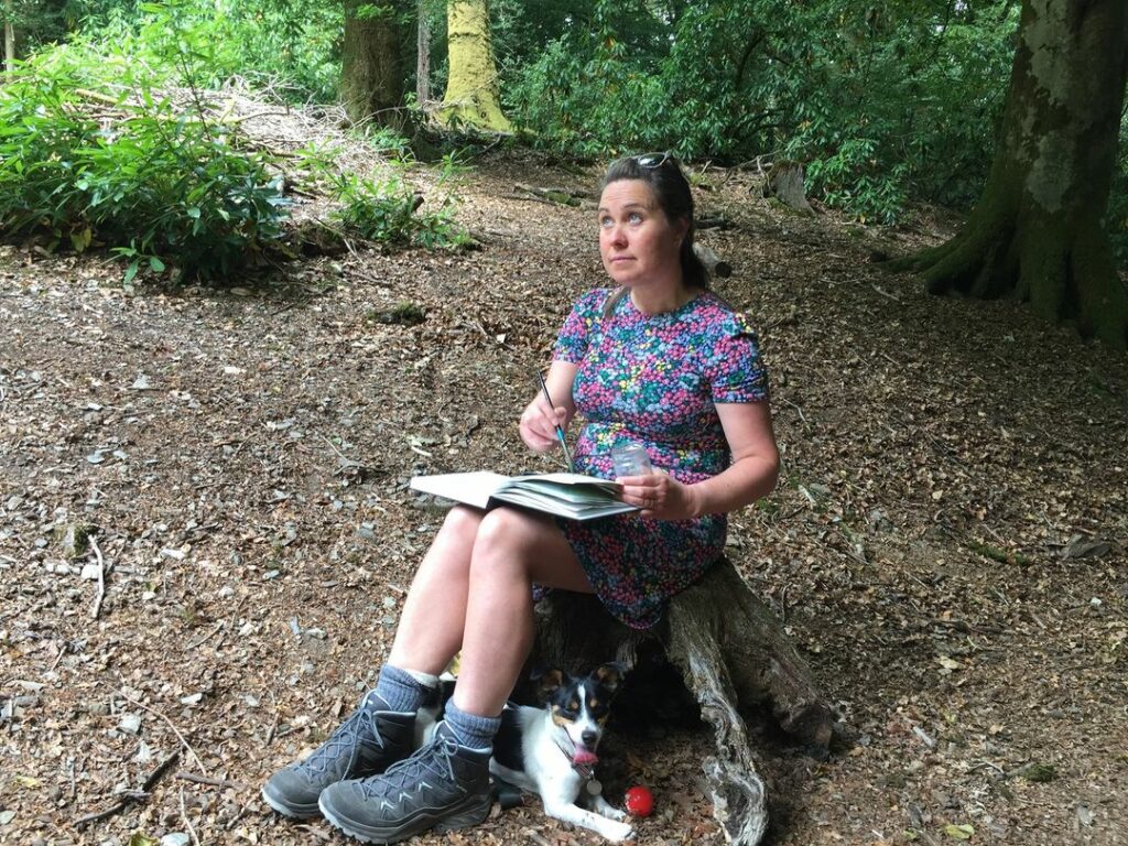 The photo depicts Felicity sat in the woods drawing. A dog is present playing with a red ball.