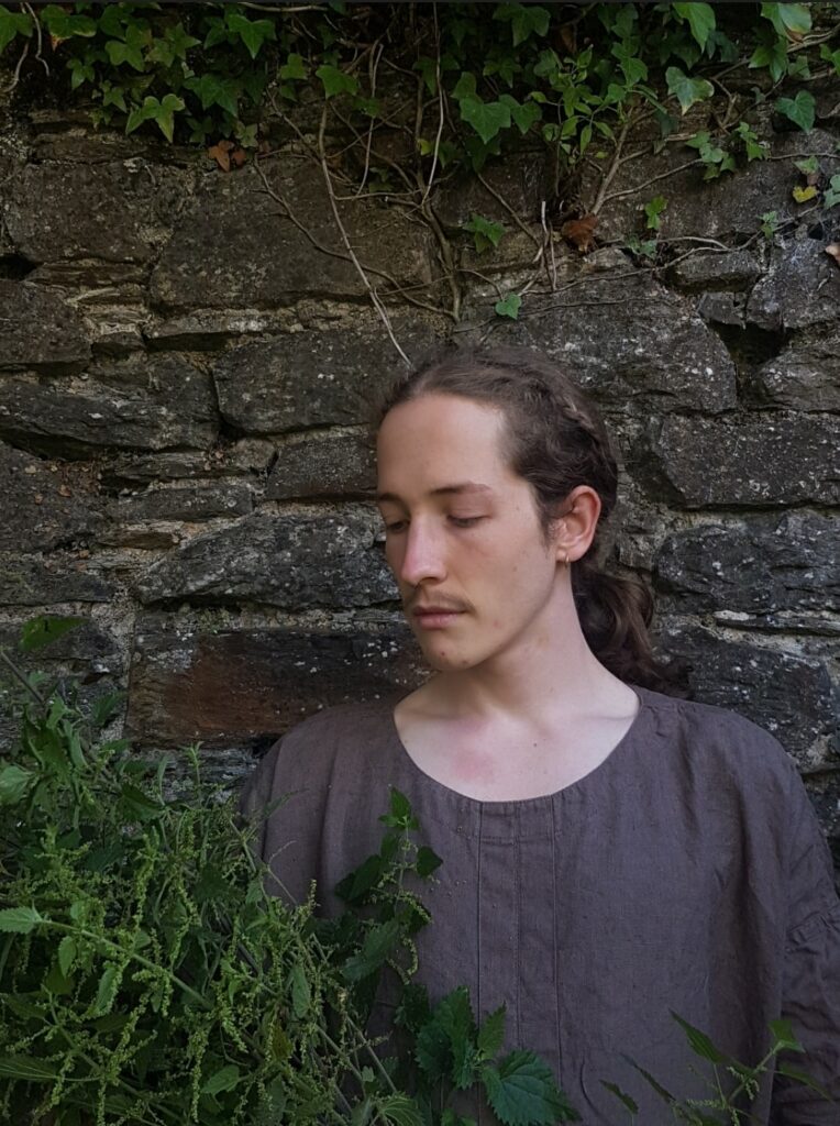 A colour photo showing artist Layan Harman in front of an old brick wall. Layan is looking at nettles while ivy climbs the top of the wall.