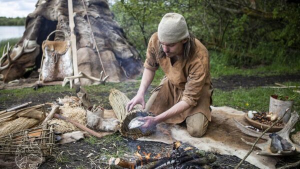man dressed in a brown smock lighting something with a campfire