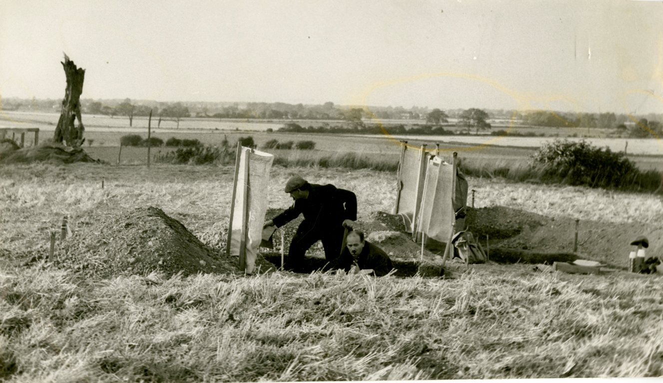 grey and white image of a man standing n a hole, countryside view behind