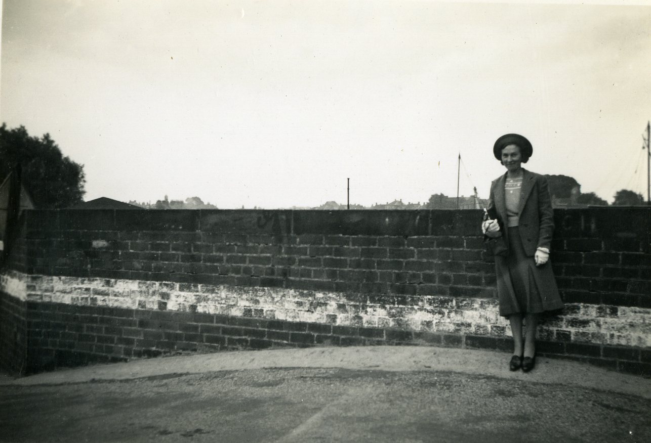 Black and white photograph of a woman wearing a hat and skirt suit, stood in front of a wall.