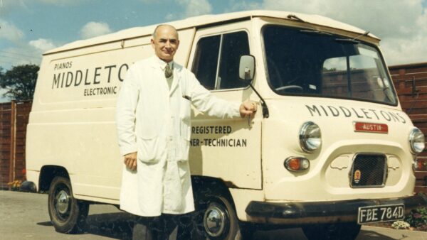 Colour photograph of a man in a white overcoat stood next to a white milk van.