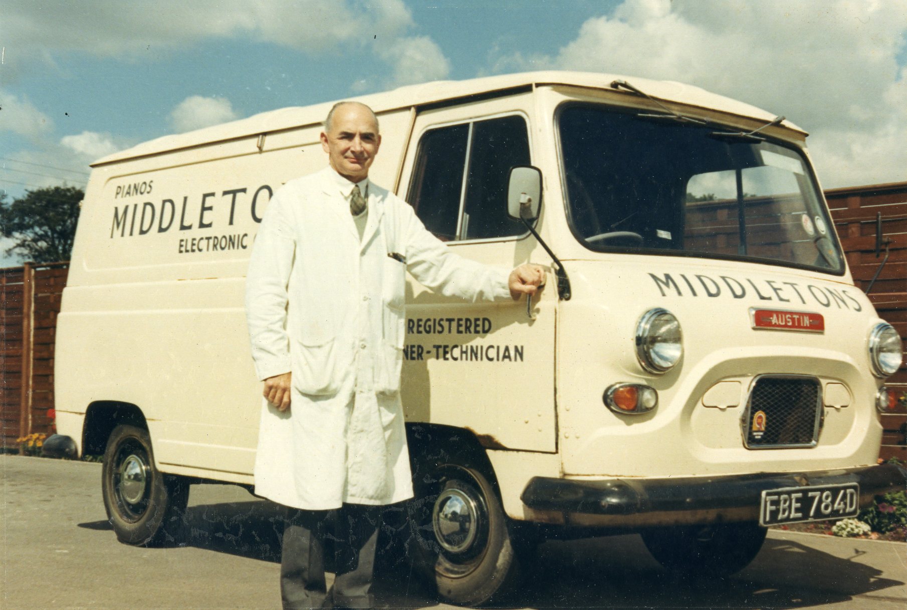 Colour photograph of a man in a white overcoat stood next to a white milk van.