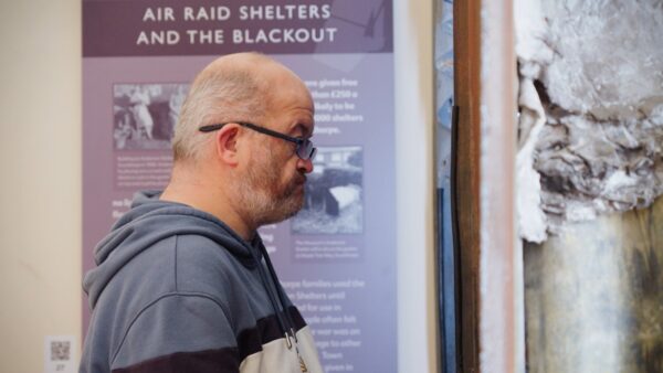 A man inside a museum gallery wearing a jumper and glasses is looking at a steel air raid shelter.