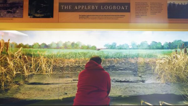 A young man in an orange jumper looks at a wooden logboat in a museum gallery.