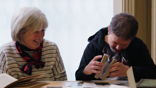 A lady with grey hair in a bob and a stripey t-shirt, sits alongside a man with a beard wearing a hoodie, sit at a table looking at documents,