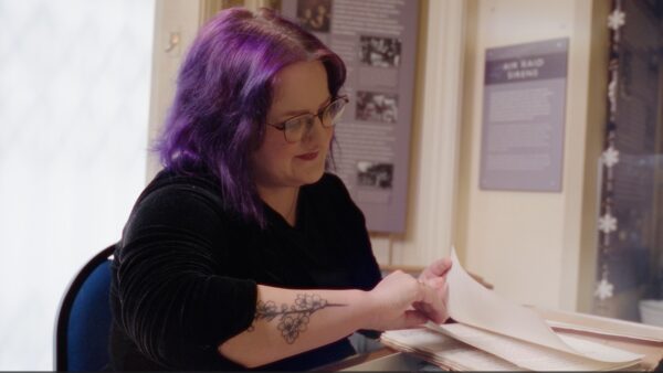 A young woman with purple hair and glasses, wearing a black top, looks at papers in a museum gallery.