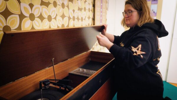 A young woman with bobbed hair and wearing a black jumper kneels next to a wooden stereogram, holing up its lid.