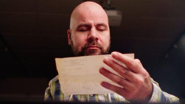A man with a bead, wearing a check shirt, holds up a document to look at it.