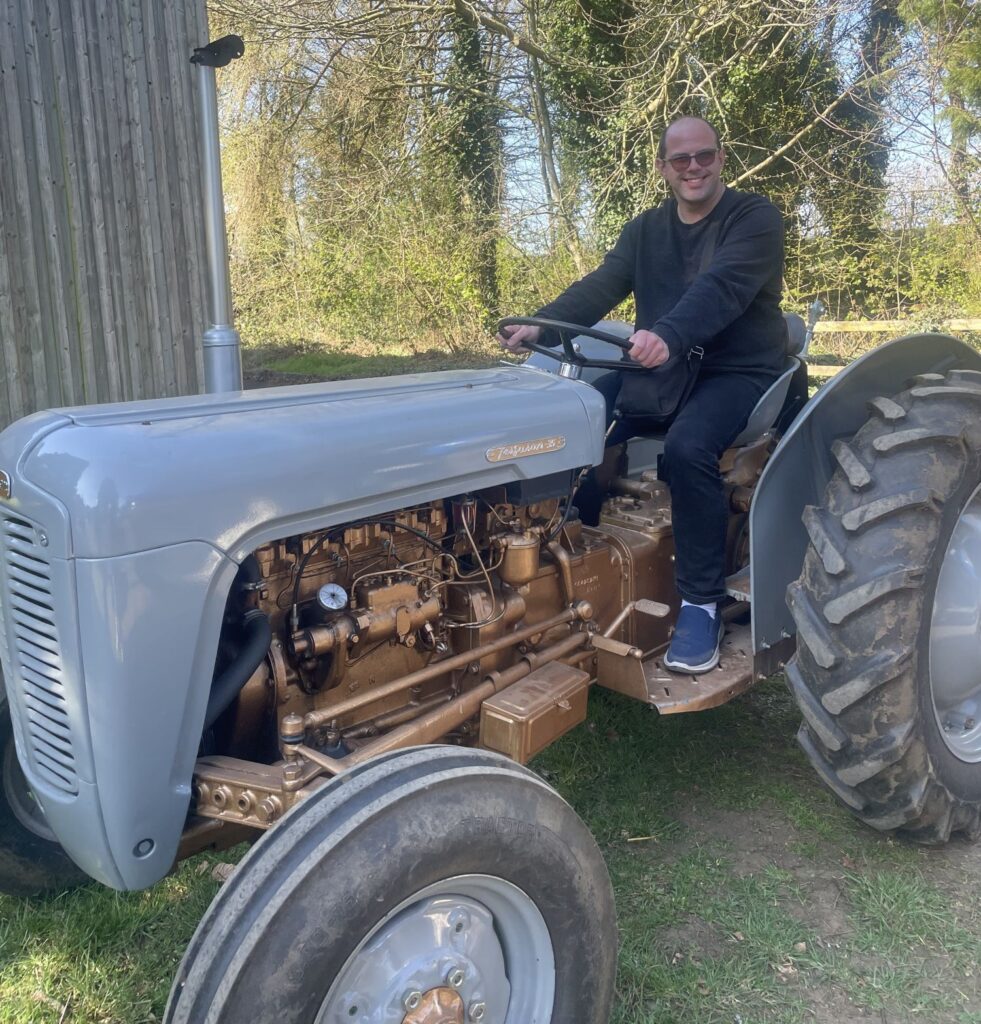 A man wearing dark clothing and glasses is sat on an old grey Massey Ferguson 35 tractor.